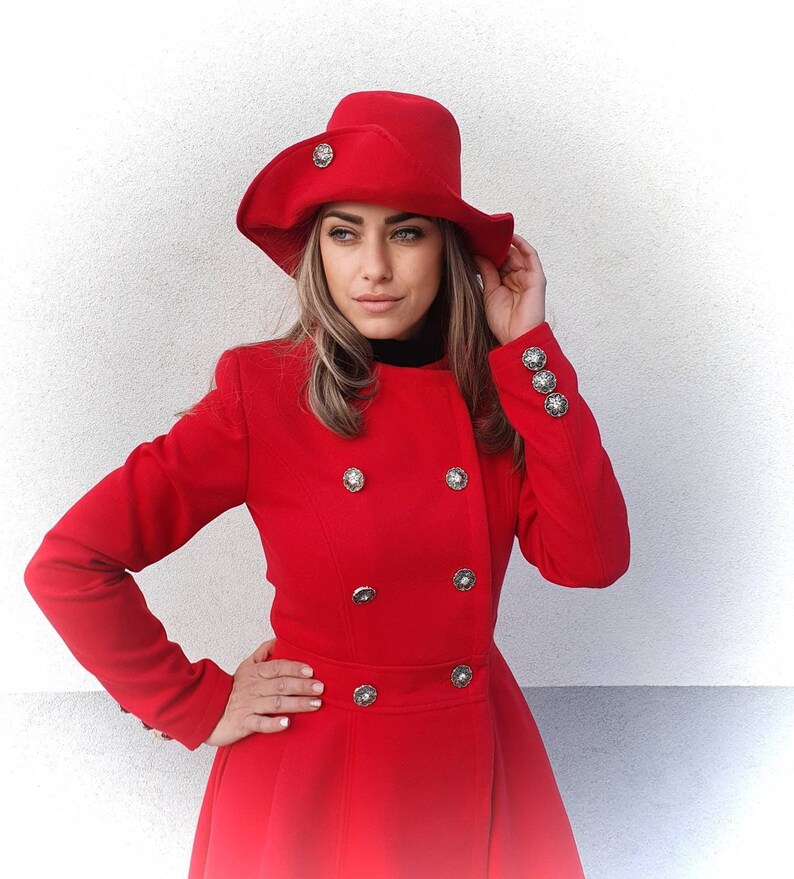 Puede incluir: Un abrigo rojo de doble botonadura con botones plateados y un sombrero rojo a juego. El abrigo tiene una falda acampanada y un cuello alto.