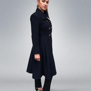May include: A woman wearing a long, dark blue coat with a high collar and gold buttons. The coat has a flared skirt and the woman is wearing black pants and heels.