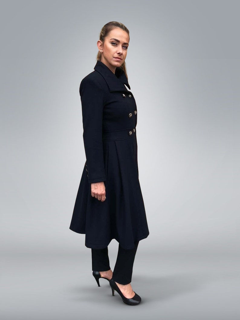 May include: A woman wearing a long, dark blue coat with a high collar and gold buttons. The coat has a flared skirt and the woman is wearing black pants and heels.