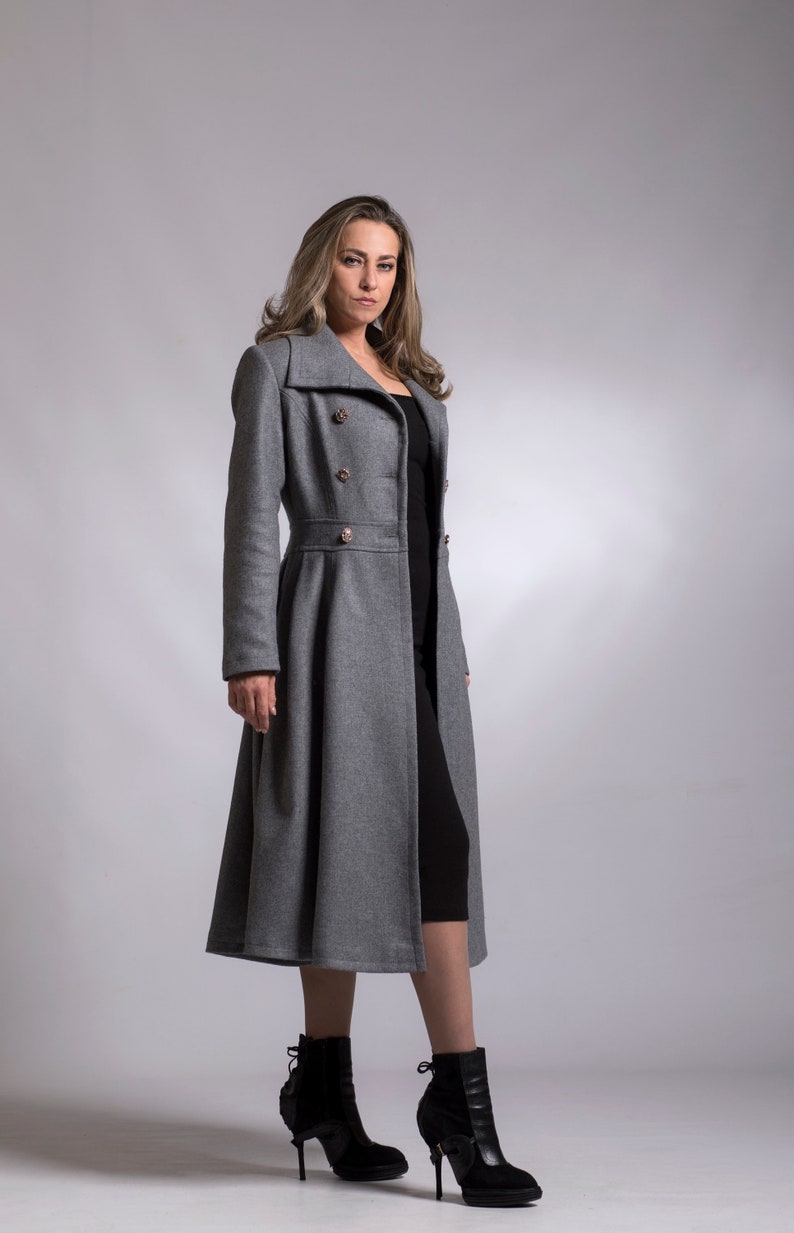 May include: A gray long coat with a double breasted closure and gold buttons. The coat has a fitted waist and a flared skirt. The model is wearing black boots.