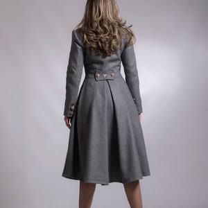 May include: A gray wool coat with a fitted bodice and a flared skirt. The coat has a high collar, long sleeves, and gold buttons. The model is wearing black heeled boots.