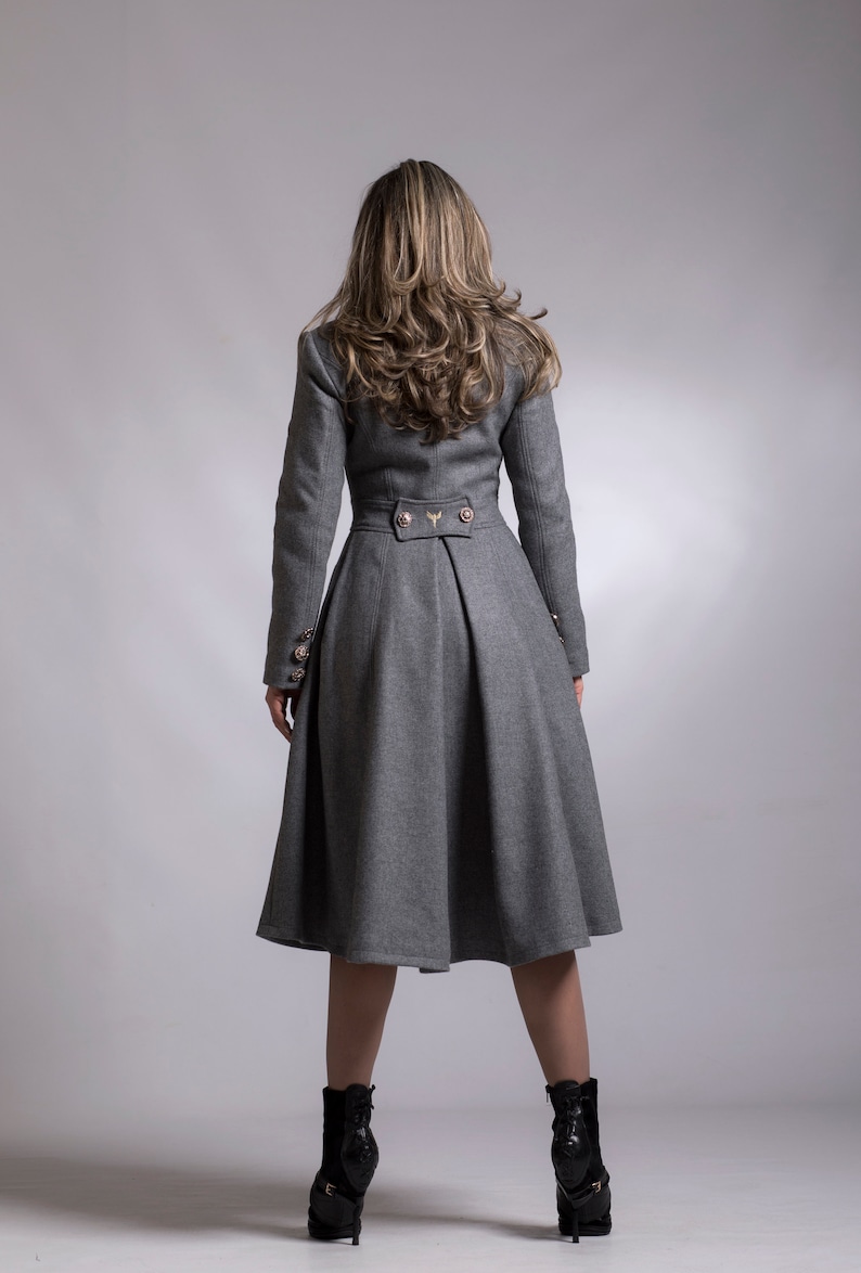 May include: A gray wool coat with a fitted bodice and a flared skirt. The coat has a high collar, long sleeves, and gold buttons. The model is wearing black heeled boots.