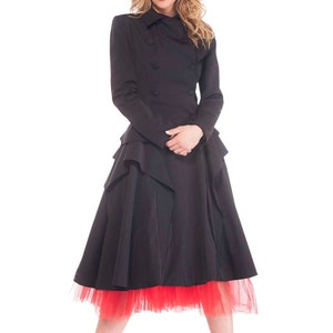 May include: A woman wearing a black double-breasted coat with a ruffled skirt and a red tulle underskirt. She is wearing red high heels.