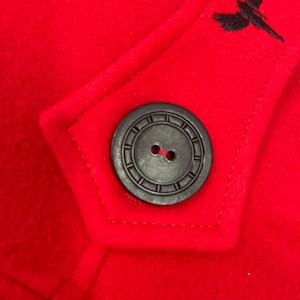 May include: A close-up of a red wool coat with a black button and a black embroidered bird design.