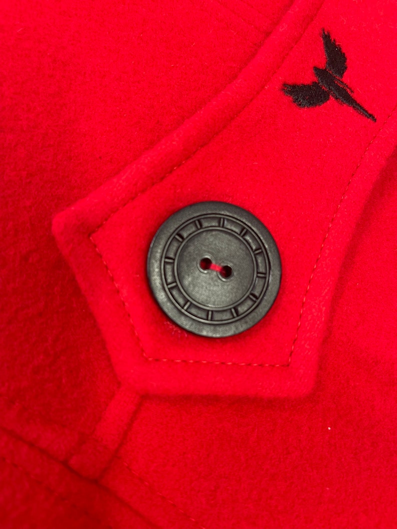 May include: A close-up of a red wool coat with a black button and a black embroidered bird design.