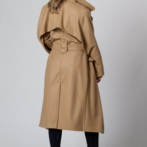 May include: A long, camel-colored coat with a belt and a large collar. The coat has a single button closure at the waist and a small embroidered design on the back.