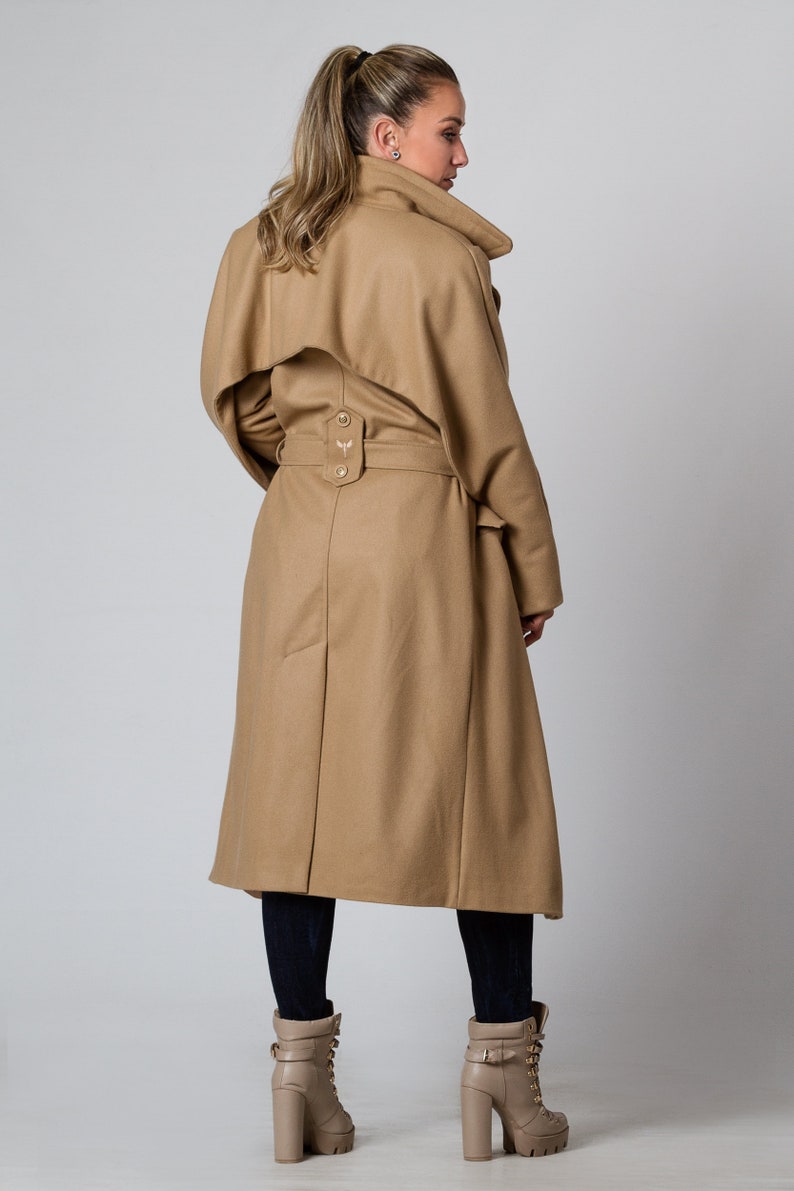 May include: A long, camel-colored coat with a belt and a large collar. The coat has a single button closure at the waist and a small embroidered design on the back.