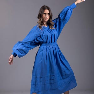 Puede incluir: Una mujer con un vestido largo de algodón azul con cuello de volantes y mangas largas. Lleva zapatillas blancas y el pelo suelto.