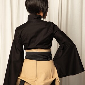 Op de afbeelding: Een zwarte top met lange, uitlopende mouwen en een hoge kraag, gecombineerd met een beige rok en een zwarte riem. De outfit is een moderne interpretatie van een klassieke stijl, met een focus op strakke lijnen en gedurfde vormen.