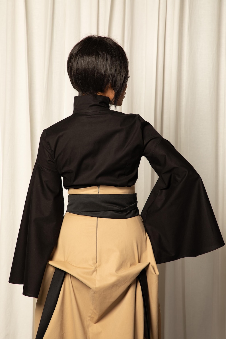 Op de afbeelding: Een zwarte top met lange, uitlopende mouwen en een hoge kraag, gecombineerd met een beige rok en een zwarte riem. De outfit is een moderne interpretatie van een klassieke stijl, met een focus op strakke lijnen en gedurfde vormen.