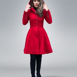 May include: A vibrant red hooded coat with silver button details and a flared skirt. The coat features long sleeves with silver buttons at the cuffs. The person is wearing black pants and black sneakers.