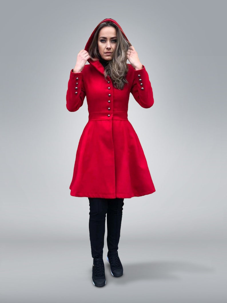 May include: A vibrant red hooded coat with silver button details and a flared skirt. The coat features long sleeves with silver buttons at the cuffs. The person is wearing black pants and black sneakers.