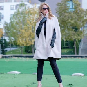 May include: A woman wearing a white cape with black leather gloves and black pants. She is standing on a green grassy surface with a building in the background.