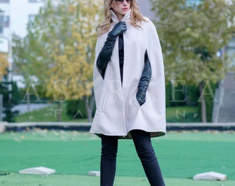 Pode incluir: Uma mulher vestindo um casaco branco com luvas de couro preto e calças pretas. Ela está em pé em uma superfície verde gramada com um prédio ao fundo.