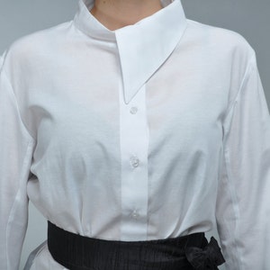 May include: A white long-sleeve shirt with a pointed collar and a black belt. The shirt has a button-up front.