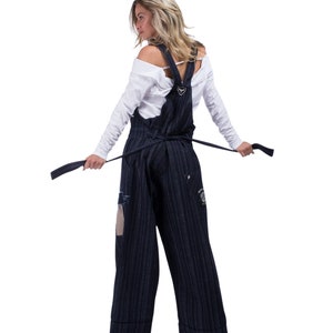 Może przedstawiać: Kobieta w białej koszuli i ciemnoniebieskich, paskowanych ogrodniczkach z sercowatą klamrą. Ogrodniczki mają przetarty wygląd i są związane w talii czarną wstążką.