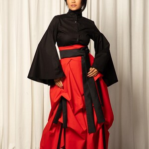 Op de afbeelding: Een zwarte top met uitlopende mouwen en een hoge kraag, gecombineerd met een rode rok. De rok is geplooid en heeft zwarte sjerpdetails. De outfit wordt afgemaakt met rode hakken.