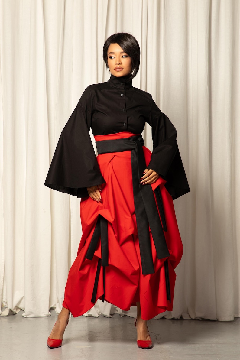 Op de afbeelding: Een zwarte top met uitlopende mouwen en een hoge kraag, gecombineerd met een rode rok. De rok is geplooid en heeft zwarte sjerpdetails. De outfit wordt afgemaakt met rode hakken.