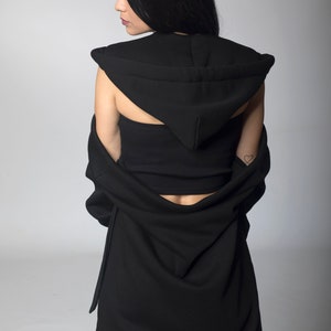Puede incluir: Vestido negro con capucha y top a juego. El vestido presenta un dise&ntilde;o drapeado y una capucha. La modelo lleva el vestido con la capucha puesta. El fondo es gris claro liso.