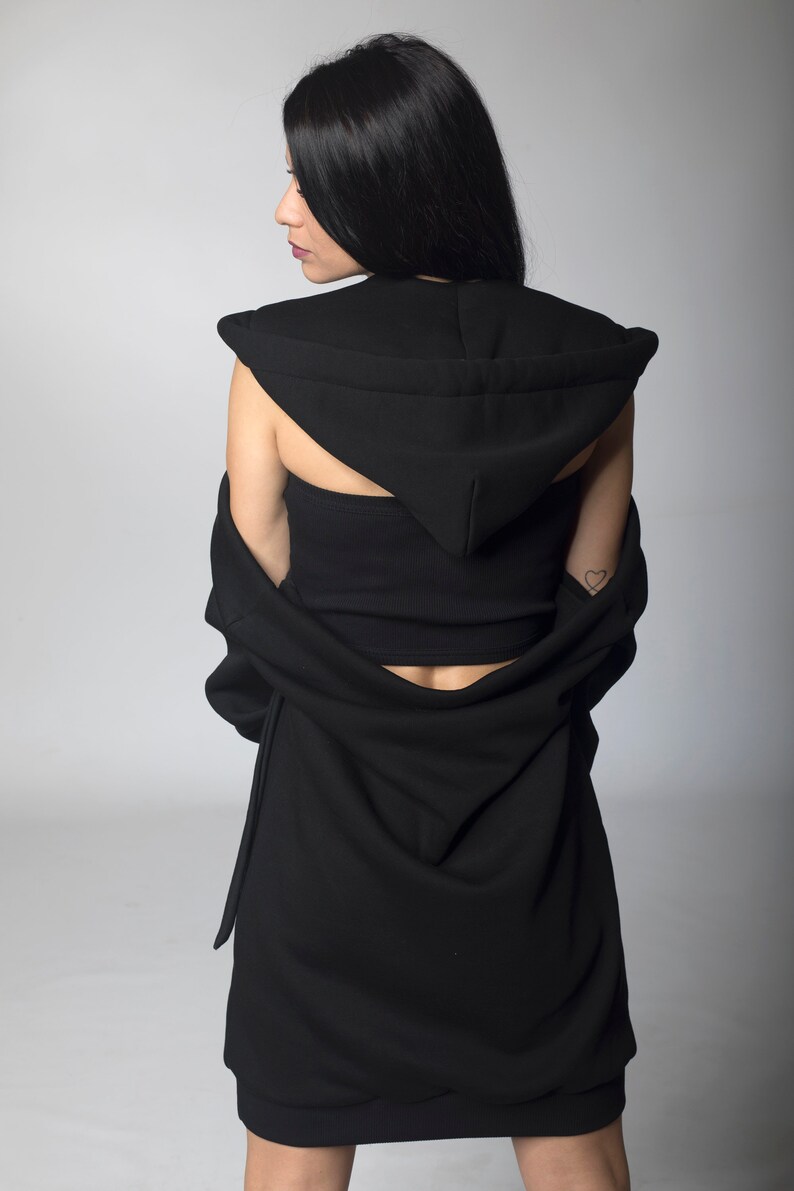 Puede incluir: Vestido negro con capucha y top a juego. El vestido presenta un dise&ntilde;o drapeado y una capucha. La modelo lleva el vestido con la capucha puesta. El fondo es gris claro liso.