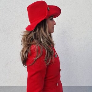Puede incluir: Una mujer con un abrigo rojo y un sombrero rojo de ala ancha con un bot&oacute;n dorado. El sombrero tiene un ala ancha que se curva hacia abajo en los lados.