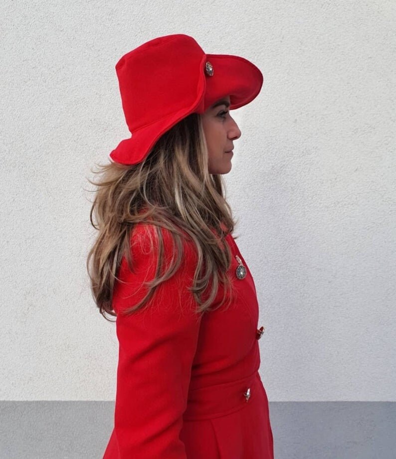 Puede incluir: Una mujer con un abrigo rojo y un sombrero rojo de ala ancha con un bot&oacute;n dorado. El sombrero tiene un ala ancha que se curva hacia abajo en los lados.
