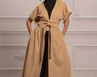 May include: A long, tan coat with a deep V-neckline and a self-tie belt. The coat has a flowing skirt and short sleeves. The model is wearing the coat with a black turtleneck and blue jeans.