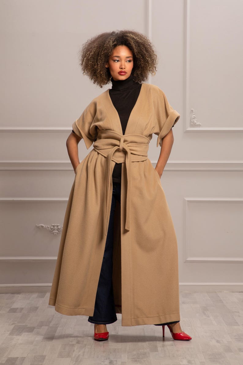 May include: A long, tan coat with a deep V-neckline and a self-tie belt. The coat has a flowing skirt and short sleeves. The model is wearing the coat with a black turtleneck and blue jeans.