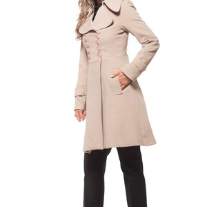 Op de afbeelding: Een beige dubbelrijdige jas met een brede kraag en lange mouwen. De jas heeft een getailleerde taille en een uitlopende rok. Het model draagt een zwarte broek en zwarte schoenen.