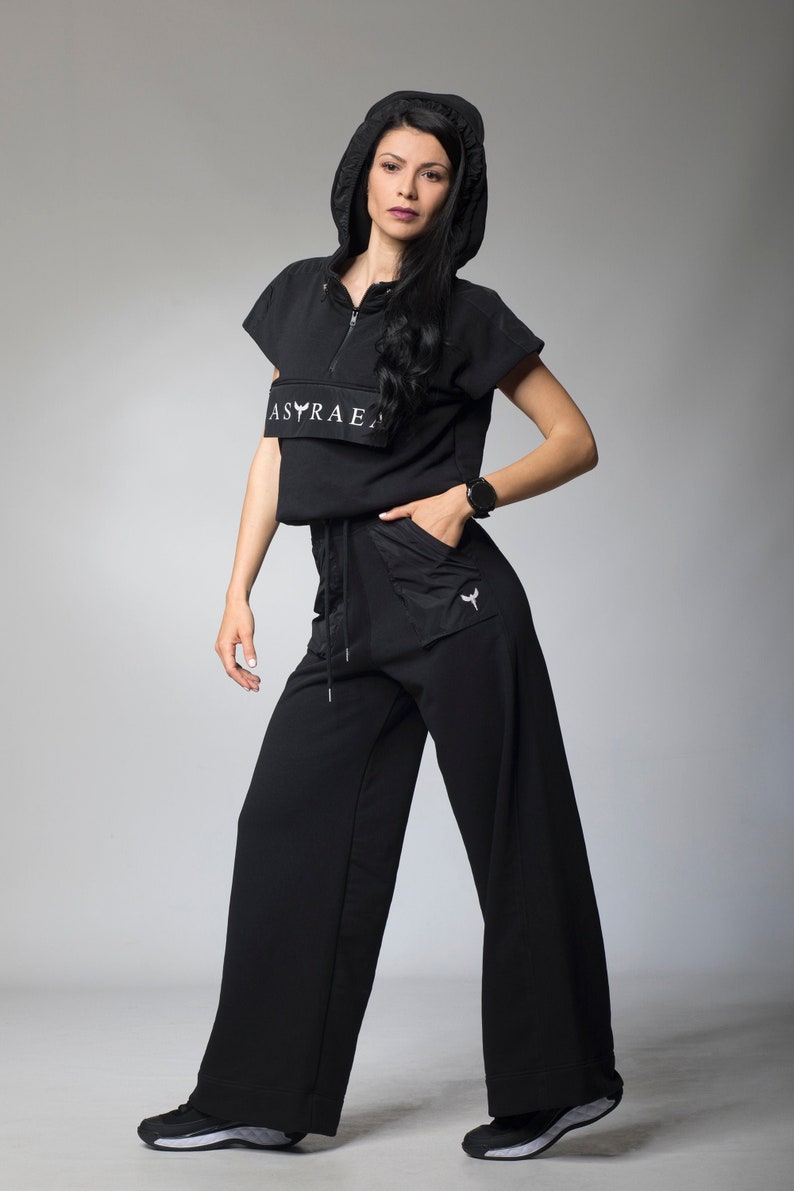 Puede incluir: Una sudadera con capucha negra de manga corta con el texto "ASRAEA" en la parte delantera. La modelo lleva pantalones negros de pierna ancha con un logotipo blanco en el bolsillo.