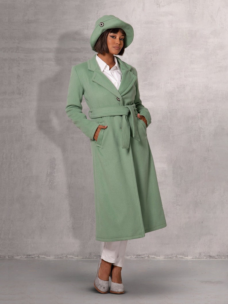 May include: A long, sage green coat with a belt and a single button fastening. The coat has a classic collar and pockets. The model is wearing a white shirt and white trousers.