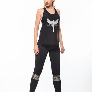May include: A black tank top with a silver graphic of a woman with wings and the text "ASTHRA" on the front. The model is wearing black leggings with silver bands on the legs that say "ASTHRA".