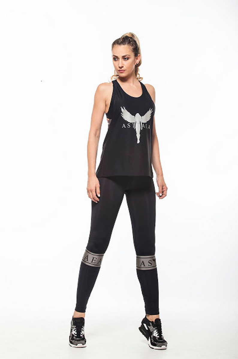May include: A black tank top with a silver graphic of a woman with wings and the text "ASTHRA" on the front. The model is wearing black leggings with silver bands on the legs that say "ASTHRA".