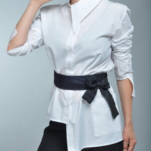 May include: A white button-down shirt with a black fabric belt tied in a bow at the front. The shirt has a collar and long sleeves.