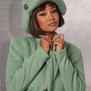 May include: A woman wearing a sage green coat with a matching sage green hat. The coat has a belt and a button fastening. The hat has a small silver button on the side.