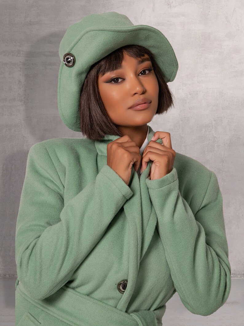 May include: A woman wearing a sage green coat with a matching sage green hat. The coat has a belt and a button fastening. The hat has a small silver button on the side.