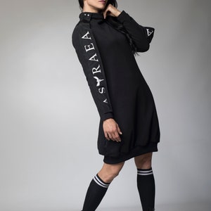 May include: A black long-sleeved dress with a mock neck and a white logo on the left sleeve that reads "ASRAEA". The dress is worn with black knee-high socks and black lace-up boots.