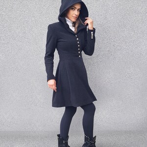 May include: A dark gray hooded coat with a flared skirt, featuring gold buttons down the front and on the sleeves. The coat is paired with black leggings and black boots with silver accents. The coat has a high collar and a fitted waist.