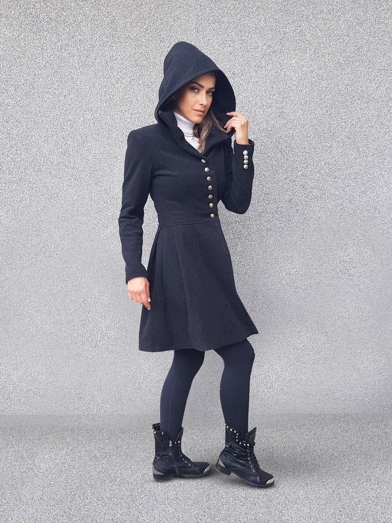 May include: A dark gray hooded coat with a flared skirt, featuring gold buttons down the front and on the sleeves. The coat is paired with black leggings and black boots with silver accents. The coat has a high collar and a fitted waist.