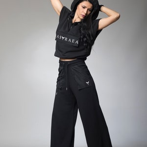 Puede incluir: Un top corto negro con capucha con el texto "ASTRAEA" impreso en la parte delantera, combinado con pantalones negros de pierna ancha. Los pantalones tienen una cintura con cord&oacute;n y un peque&ntilde;o bolsillo en la pierna derecha.