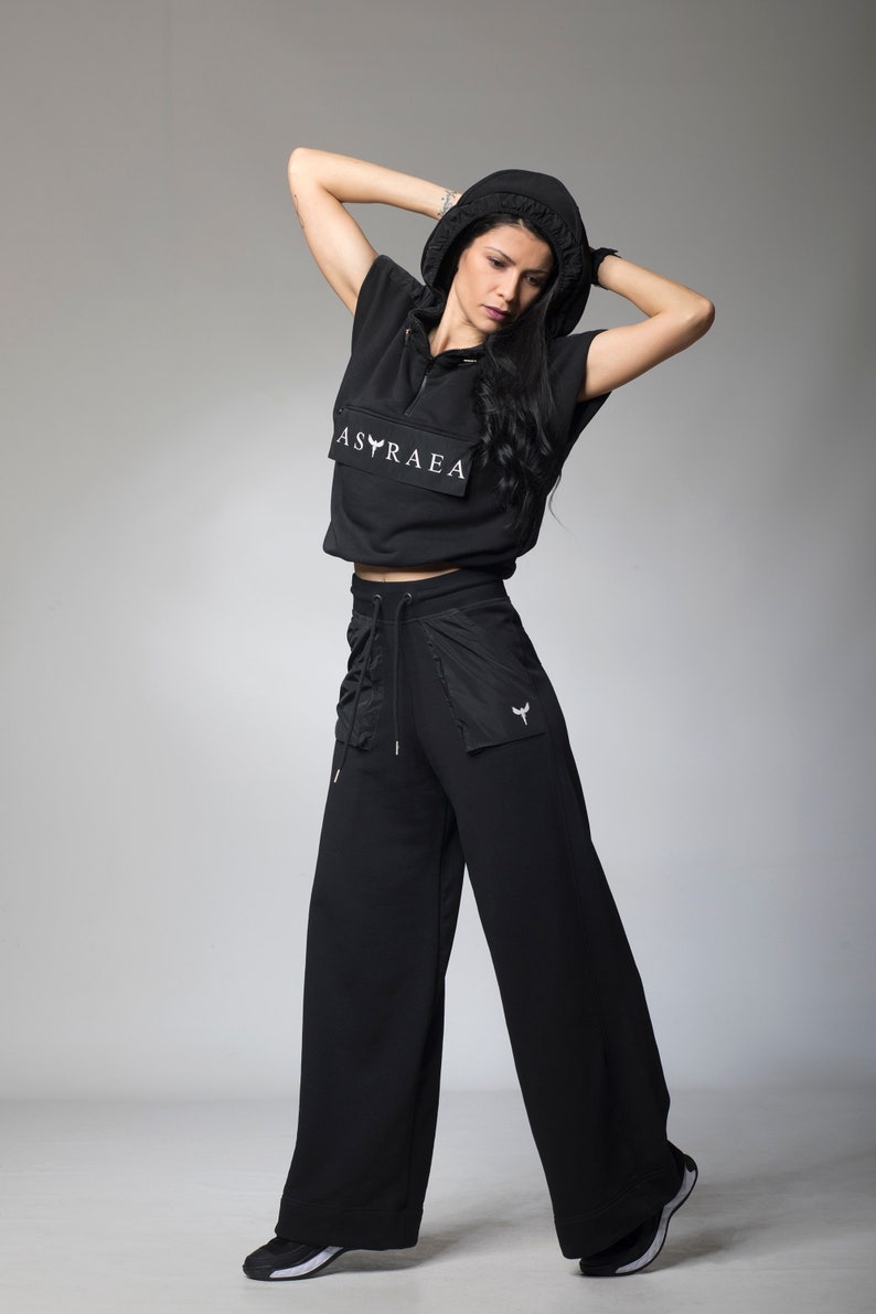 Puede incluir: Un top corto negro con capucha con el texto "ASTRAEA" impreso en la parte delantera, combinado con pantalones negros de pierna ancha. Los pantalones tienen una cintura con cord&oacute;n y un peque&ntilde;o bolsillo en la pierna derecha.