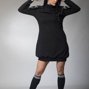 May include: A black mock neck dress with white lettering on the shoulders. The dress is worn with black knee-high socks and black platform boots.