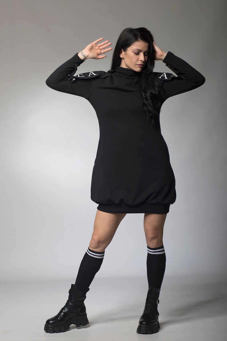 May include: A black mock neck dress with white lettering on the shoulders. The dress is worn with black knee-high socks and black platform boots.