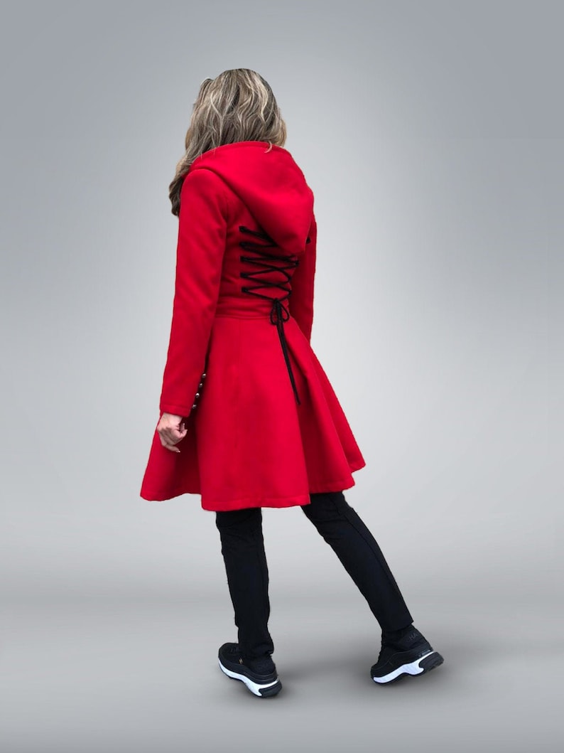 May include: A bright red hooded coat with a flared skirt and black corset-style lacing down the back. The coat features long sleeves and is paired with black leggings and sneakers.