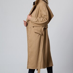 May include: A long camel-colored coat with a belt and a large pocket on the front. The coat has a collar and is made of a soft, warm fabric.