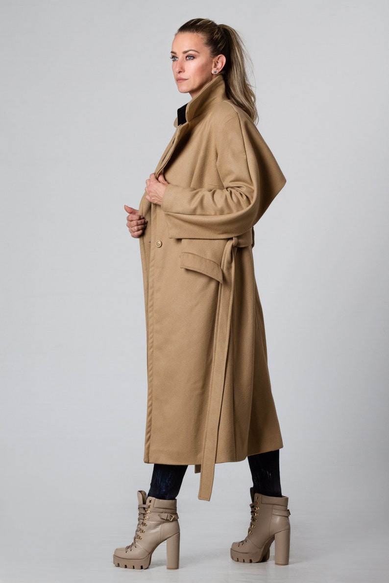 May include: A long camel-colored coat with a belt and a large pocket on the front. The coat has a collar and is made of a soft, warm fabric.