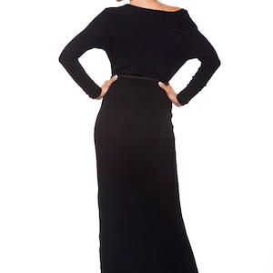 May include: A woman wearing a long black dress with a fitted waist and a slightly off-the-shoulder neckline. She is standing with her hands on her hips and looking down.