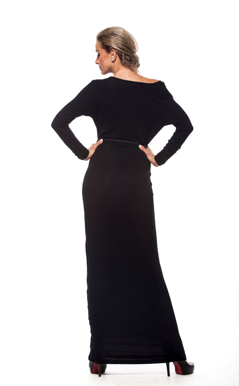 May include: A woman wearing a long black dress with a fitted waist and a slightly off-the-shoulder neckline. She is standing with her hands on her hips and looking down.