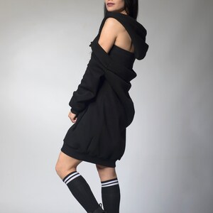 Puede incluir: Un vestido negro con capucha de dise&ntilde;o &uacute;nico, con una capucha drapeada y una longitud mini. El atuendo se combina con botas de combate negras y calcetines hasta la rodilla con rayas blancas. La modelo est&aacute; de pie frente a un fondo gris.