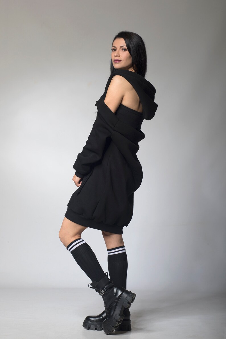 Puede incluir: Un vestido negro con capucha de dise&ntilde;o &uacute;nico, con una capucha drapeada y una longitud mini. El atuendo se combina con botas de combate negras y calcetines hasta la rodilla con rayas blancas. La modelo est&aacute; de pie frente a un fondo gris.